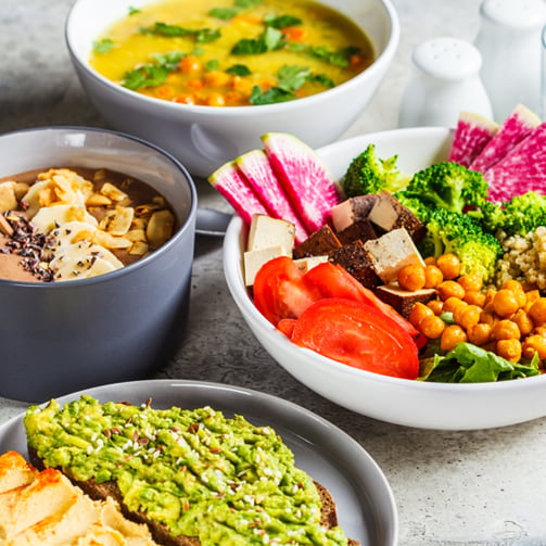same Copilot said: Top-down view of a variety of prepared dishes, including vegetable soup, a fresh salad with tofu and chickpeas, grain porridge with sliced almonds, and avocado toast, arranged on a tabletop.