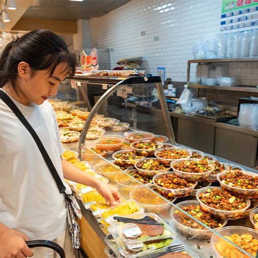 Customer selecting a packaged food item from a food display counter with prepared dishes.
