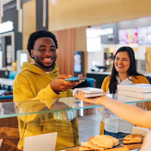 Person paying with a smartphone at a food counter while receiving a takeaway meal; another customer waits nearby.