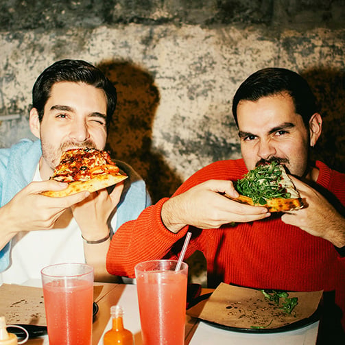 Two people seated at a table eating large slices of pizza, with colorful beverages, condiments, and plates visible in a casual indoor dining setting.