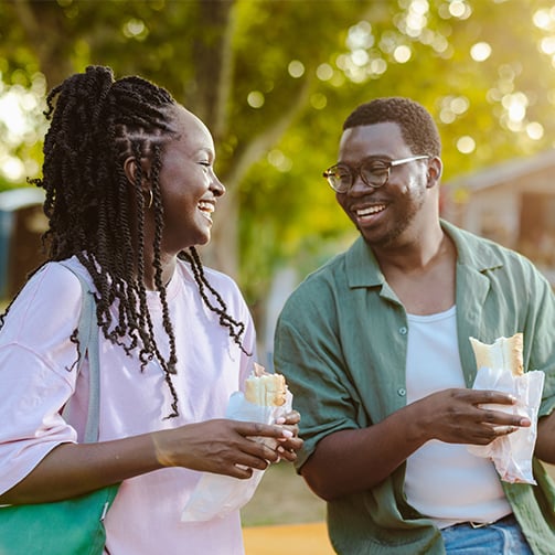 Two people outdoors talking while holding wrapped sandwiches in a sunlit park setting.