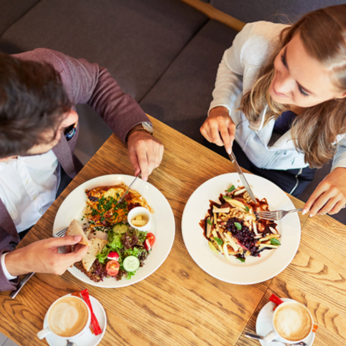 Overhead view of two people dining with plated meals and coffee at a wooden table.
