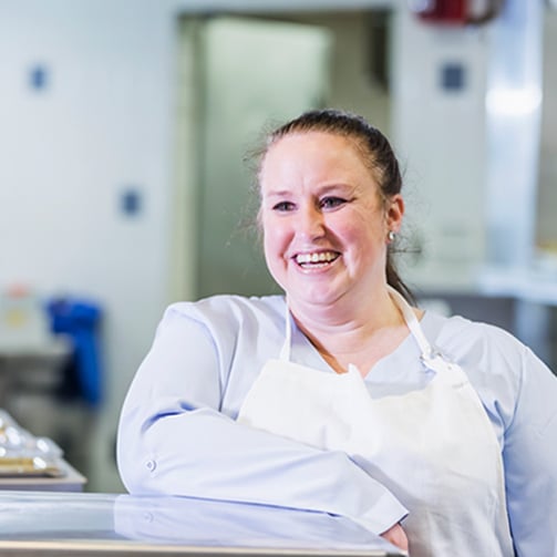 Person in white apron leaning on counter in a commercial kitchen setting.