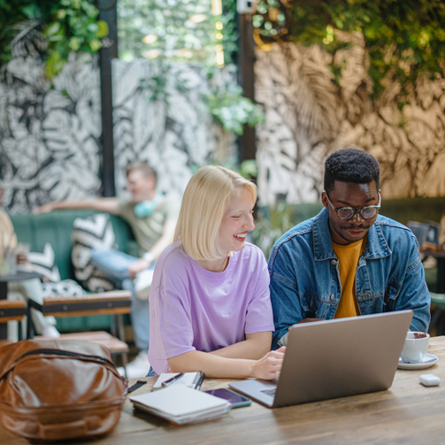 Two people sitting at a café table, working together on a laptop with notebooks and coffee nearby.