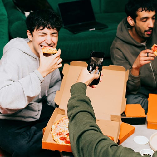 People sitting together indoors eating pizza from open delivery boxes while another person takes a photo with a smartphone, creating a casual shared dining scene.