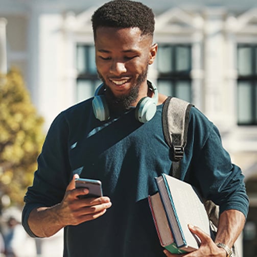 Person walking outdoors while looking at a smartphone, carrying books and wearing headphones around the neck, with a backpack and a campus-style building blurred in the background.
