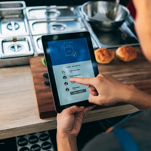 Person using a tablet to track food order status in a commercial kitchen, with progress updates displayed on screen and food preparation equipment visible on the counter nearby.