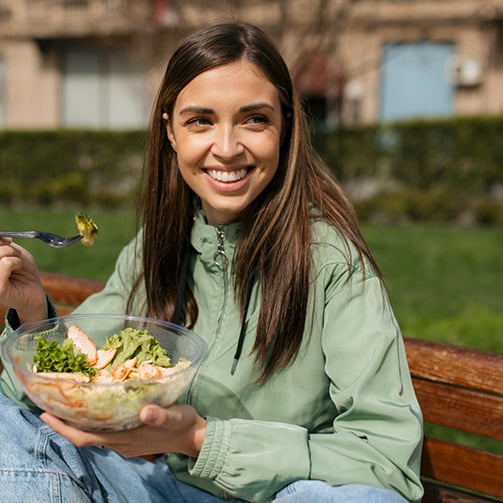 Person sitting outdoors on a wooden bench holding a bowl of salad and a fork, with leafy greens and sliced protein visible in the bowl and a campus style setting in the background.