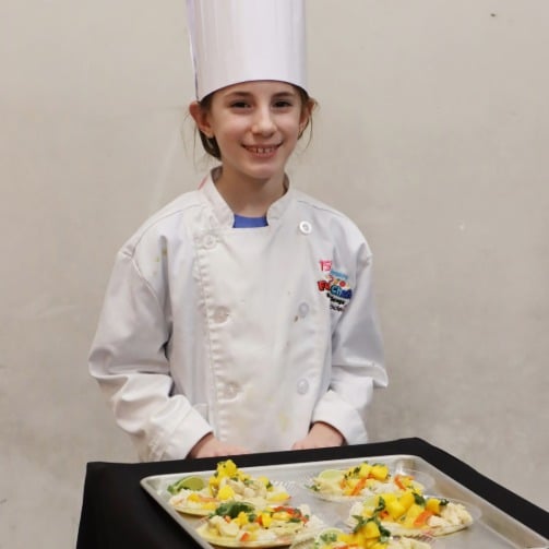 Child chef in white jacket and tall hat standing behind tray of prepared dishes on a table.