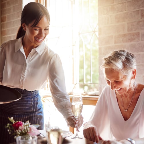 A server handing a glass of champagne to a smiling female senior resident, capturing a moment of attentive service.