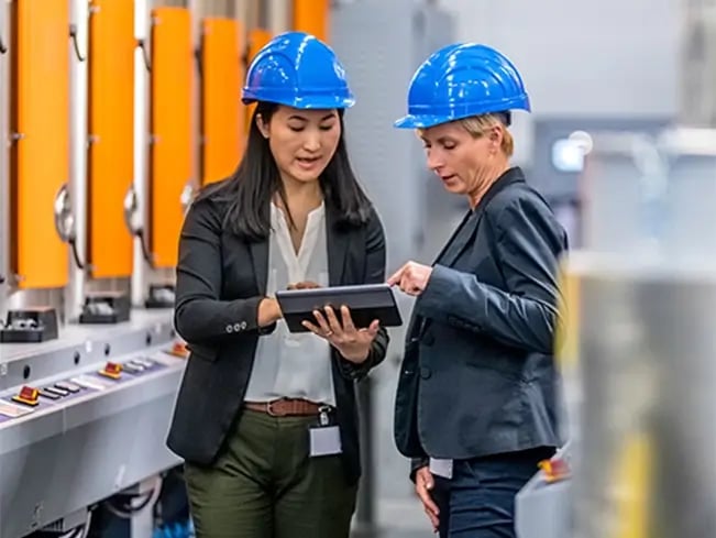 Two people in blue hard hats and business attire review a tablet by orange industrial machinery.