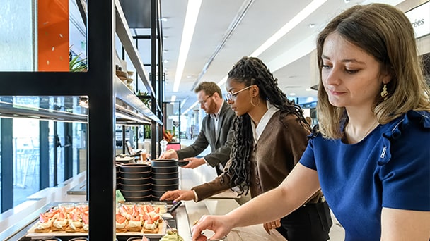 Three people serve themselves from a buffet in a modern glass-walled cafeteria.