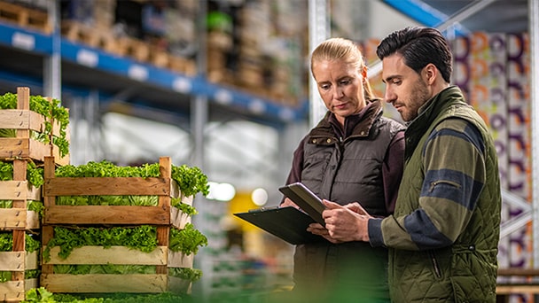 Two people in a warehouse inspect crates of fresh lettuce; one holds a clipboard, the other a tablet.