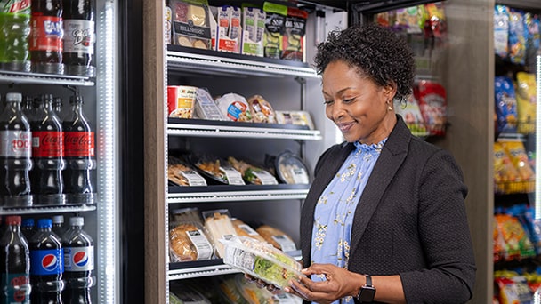 Customer picks a packaged salad from a refrigerated grocery shelf stocked with drinks and snacks.