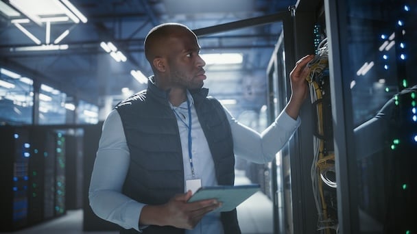 Technician holding a tablet while inspecting server rack cabling in a data center.