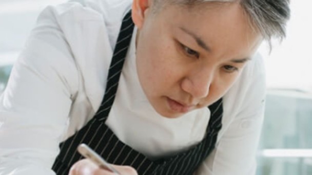 Chef in striped apron carefully plating a dish in a bright kitchen setting.