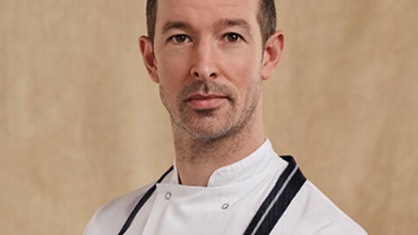 Portrait of a chef in a white jacket and apron against a neutral background.