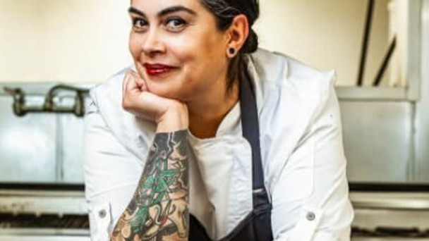 Chef in white jacket with forearm tattoos resting chin on hand in a kitchen setting.