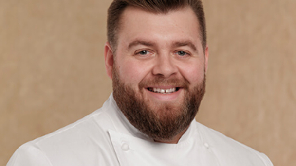Portrait of a smiling chef with a beard in a white jacket against a plain neutral background.