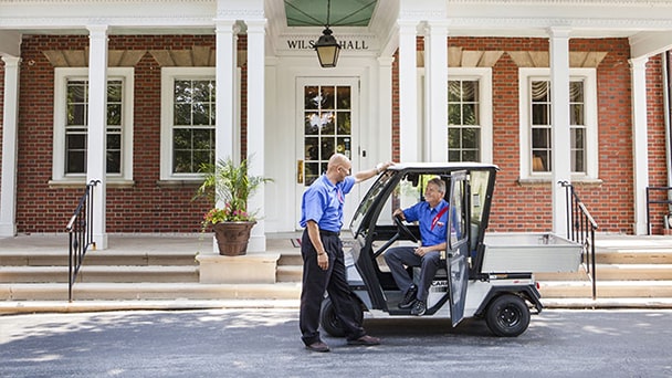 Two men sit in a golf cart outside a large brick building, enjoying a sunny day.