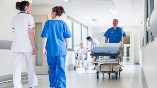 Healthcare professionals walk down a hospital corridor, with a doctor pushing an empty patient bed. 