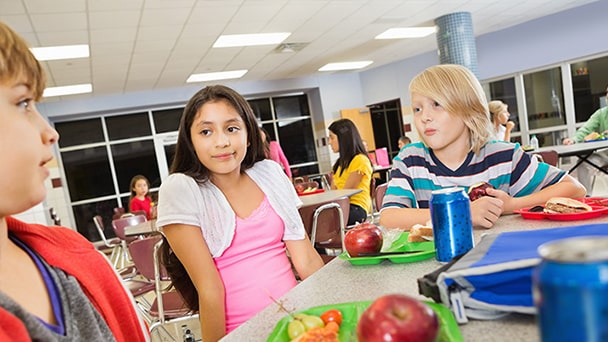 Children enjoying lunch together in a bustling cafeteria, seated at tables with trays of food.