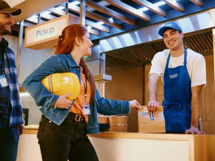 Person at a food pickup counter receiving a takeaway bag from a worker wearing an apron, while another person stands nearby holding a hard hat under a sign labeled pickup.