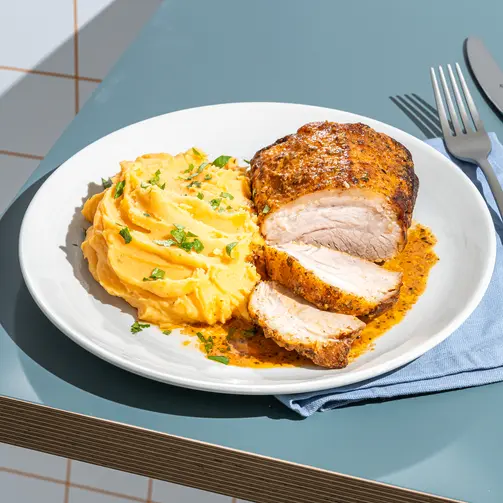Close-up of a plated meal with sliced roasted chicken breast seasoned with spices, served alongside creamy mashed potatoes garnished with herbs on a white dish over a blue table surface.