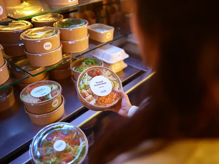 Close-up of a person selecting a packaged noodle dish from a refrigerated display filled with labeled takeaway containers, arranged on glass shelves under warm lighting.
