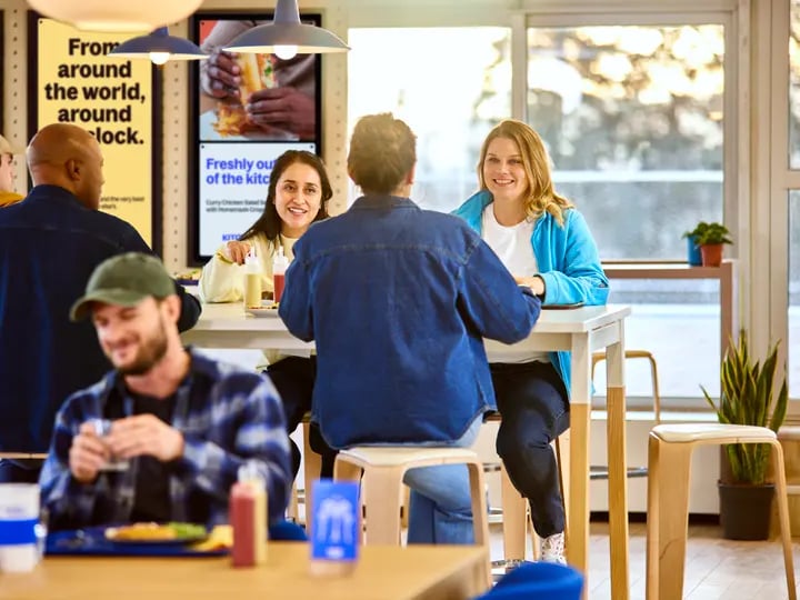 People seated around high tables in a bright cafeteria, eating and talking, with digital menu boards on the wall and large windows letting in natural light.