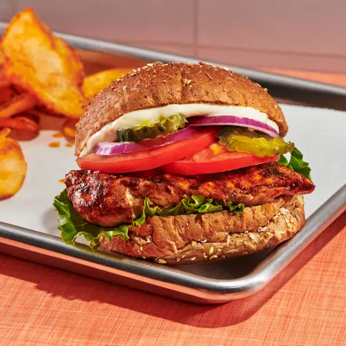 Close-up of a grilled chicken sandwich on a whole grain bun with lettuce, tomato, pickles, and red onion, served on a tray with potato chips on a warm-toned table surface.