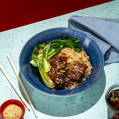 Top-down view of a bowl with rice, glazed meat pieces garnished with sesame seeds and sliced peppers, served with leafy greens, chopsticks, and small bowls on a textured table surface.