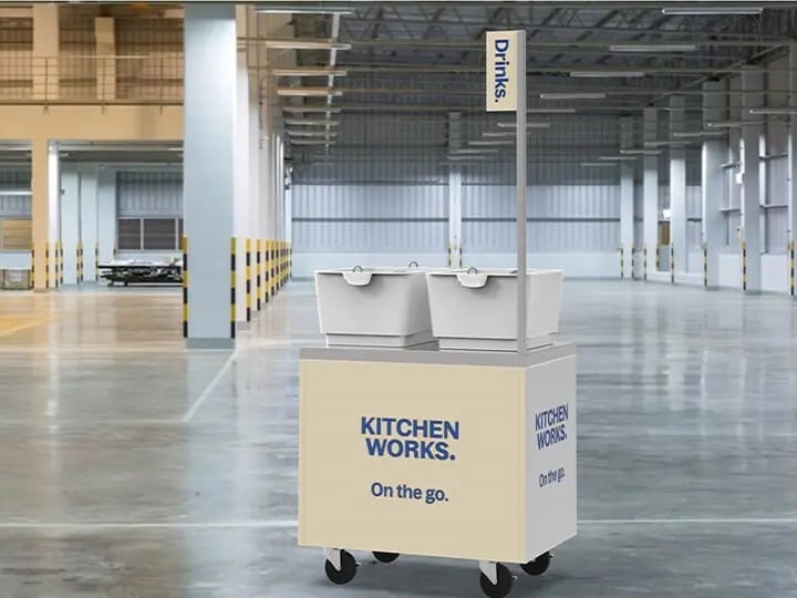 Mobile refreshment cart in a large industrial warehouse, labeled Kitchen Works On the go, with two drink containers on top and a Drinks sign above, surrounded by wide open floor space.