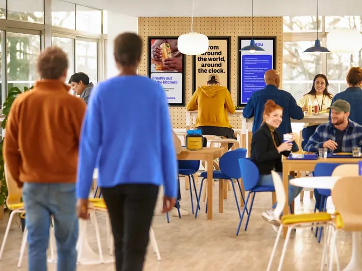 People walking and sitting in a bright cafeteria with blue chairs and wooden tables, ordering food from digital menu boards and eating together in a modern, casual dining space.