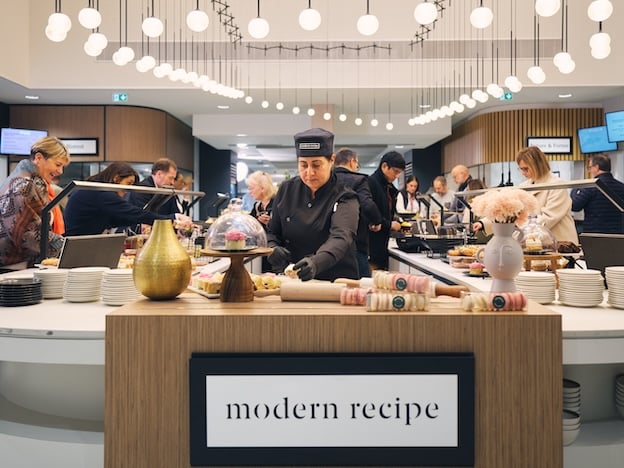 Busy modern restaurant counter as staff prepare desserts under hanging lights, with guests around and a "modern recipe" sign.