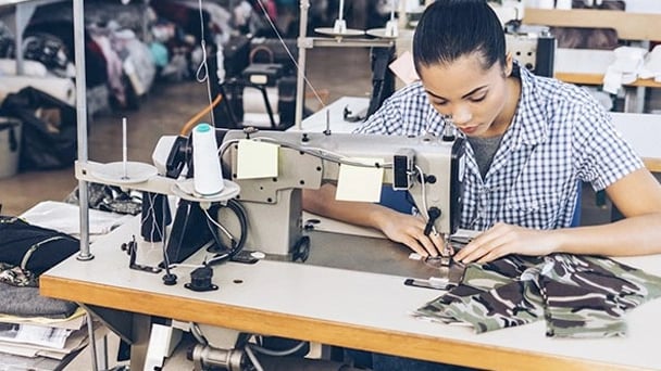 A woman operates a sewing machine in a factory, focused on her work amidst a busy industrial environment.