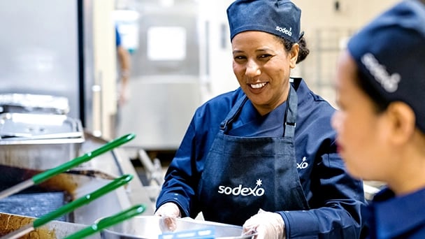 Two women in blue uniforms are preparing food together in a kitchen setting.