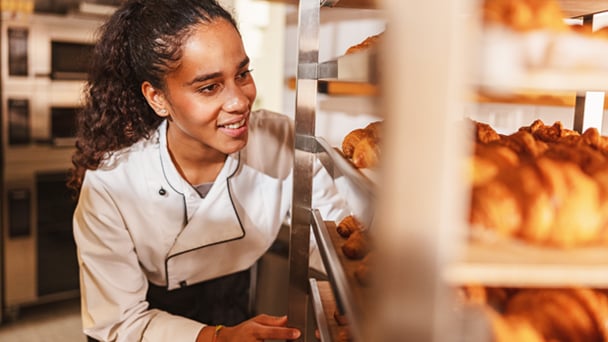 A young chef in a white uniform smiles while checking trays of freshly baked croissants in a commercial kitchen