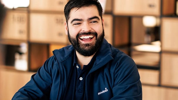 Bearded man in a navy blue Sodexo jacket sits indoors, smiling, with wooden shelves in the softly blurred background.