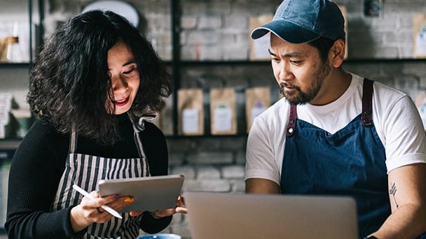 A man and woman in aprons collaborate on a laptop, focused on their work in a cozy kitchen setting.