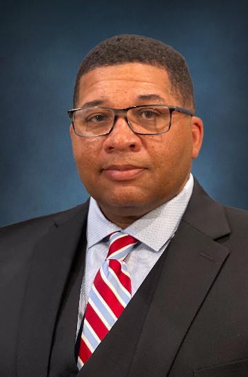 Professional headshot of a person wearing glasses, a dark suit, and a red striped tie against a blue background.