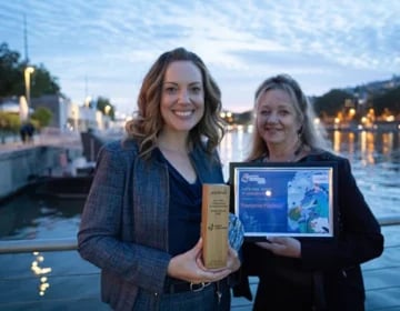 Two people holding an award and framed certificate by a waterfront during twilight.