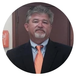 Person in black suit, blue shirt, and orange tie standing in front of a brown door.