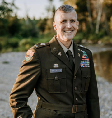 Person in military uniform with badges and insignia stands outdoors with trees in the background.