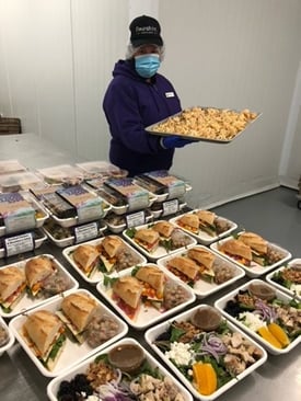 Person in purple hoodie holding food tray, with prepared meal trays on table in front.
