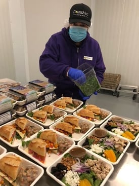 Person in purple jacket and gloves preparing meal trays with sandwiches, salads, and toppings.