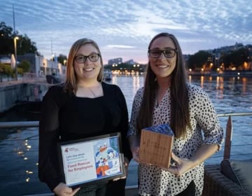 Two people holding awards by a river at dusk with a cityscape in the background.