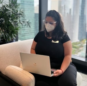 Person on beige couch using silver laptop with cityscape visible through large window behind.