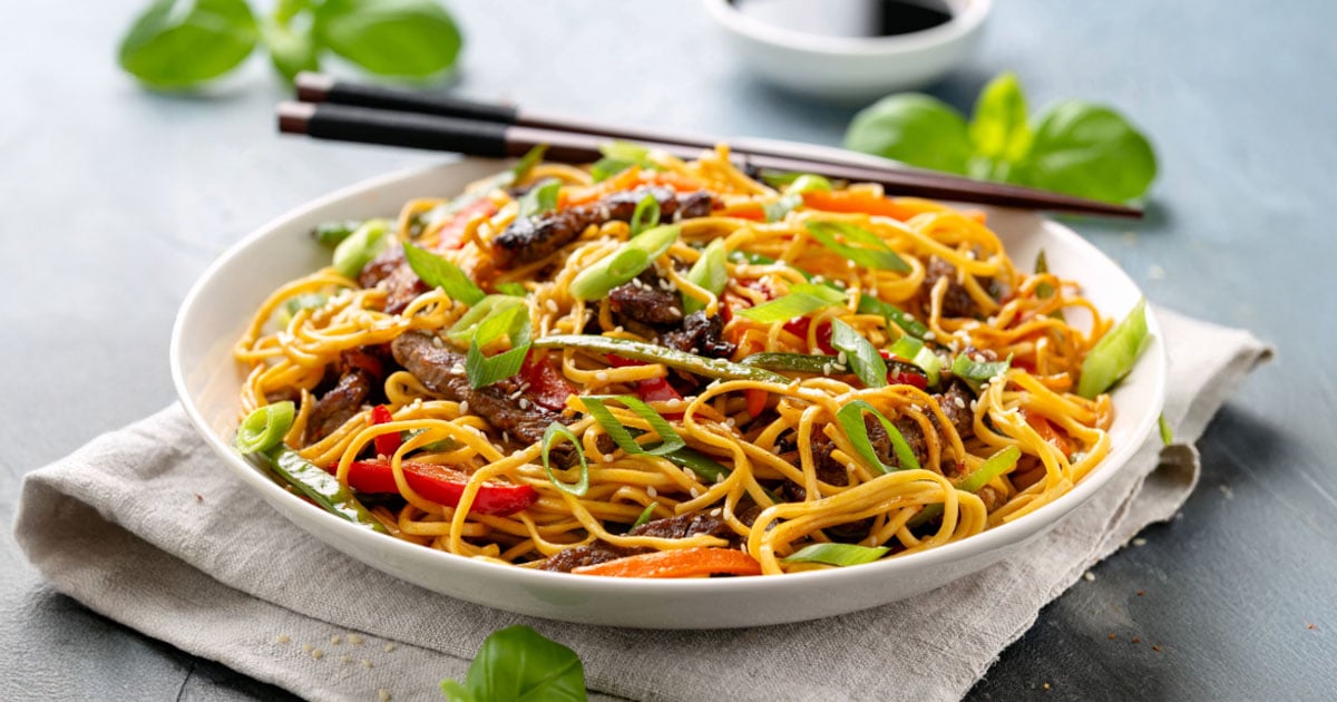 Close-up of an Asian-inspired beef stir-fry noodle bowl with vegetables and sprinkled sesame seeds. 