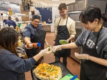Four people in a kitchen preparing food, adding sauce and sprinkling herbs on a dish.